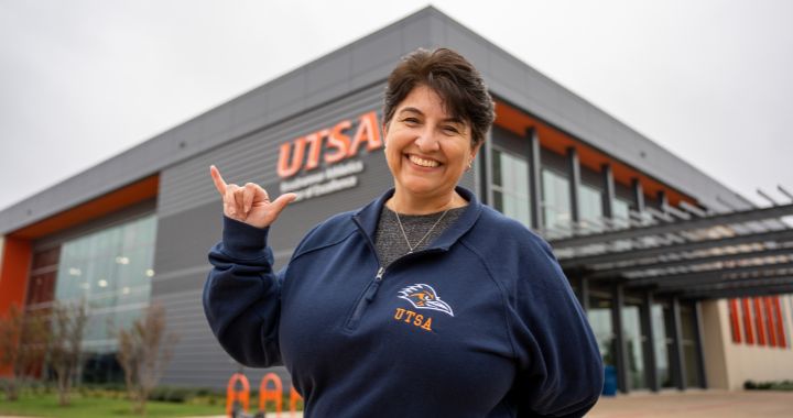 UTSA Online alum Brenda Davila holding up a Roadrunner sign while standing in front of the UTSA Race building